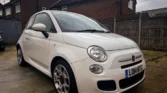 A white Fiat 500 hatchback parked on a driveway. The car appears to be in the 'S' trim level with distinctive alloy wheels. It's photographed at an angle showing the front quarter and driver's side against a backdrop of brick houses and wooden fencing on an overcast day.