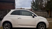 A white Fiat 500 hatchback parked on a driveway. The car appears to be in the 'S' trim level with distinctive alloy wheels. It's photographed at an angle showing the Side view against a backdrop of brick houses and wooden fencing on an overcast day.