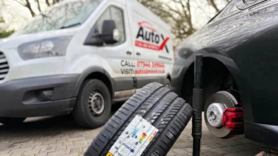 Mobile tyre fitting in progress with a car lifted on a jack, wheel removed, and new tyres stacked nearby, while an Auto X Limited service van is parked in the background on a residential driveway.